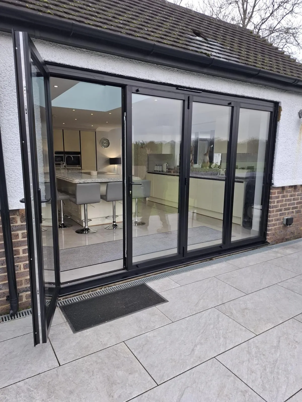 Anthracite aluminium bifold doors, one leaf open, between a kitchen and a porcelain-paved patio