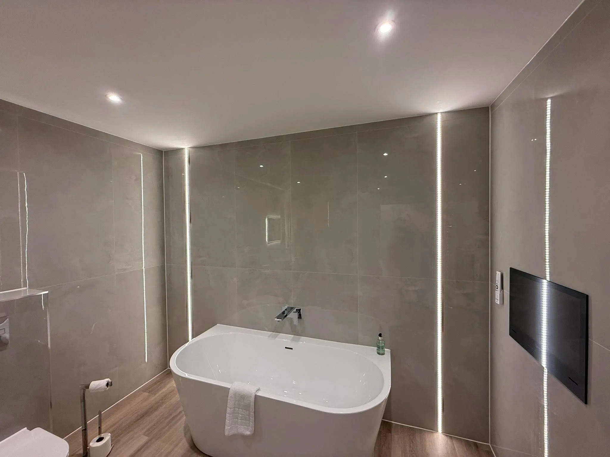 Freestanding bath against large-format grey wall tiles, with vertical LED strip lighting