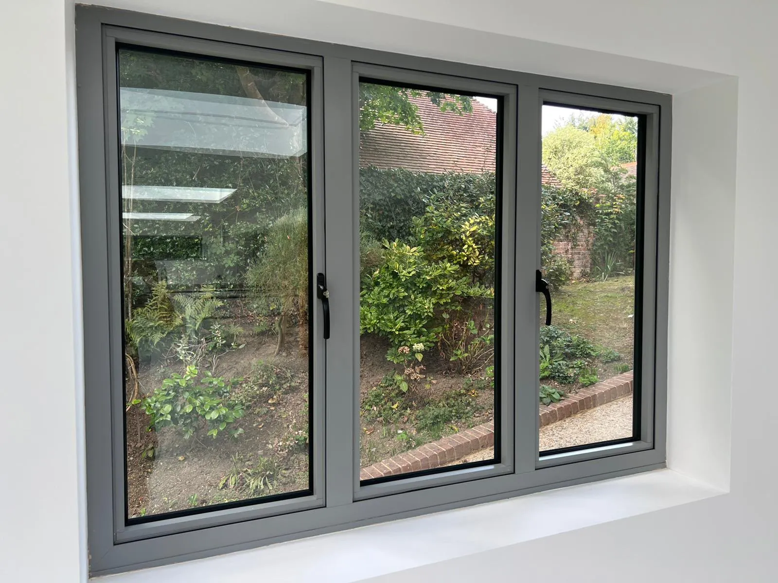 Grey aluminium three-light window with black handles, seen from inside, looking onto a garden